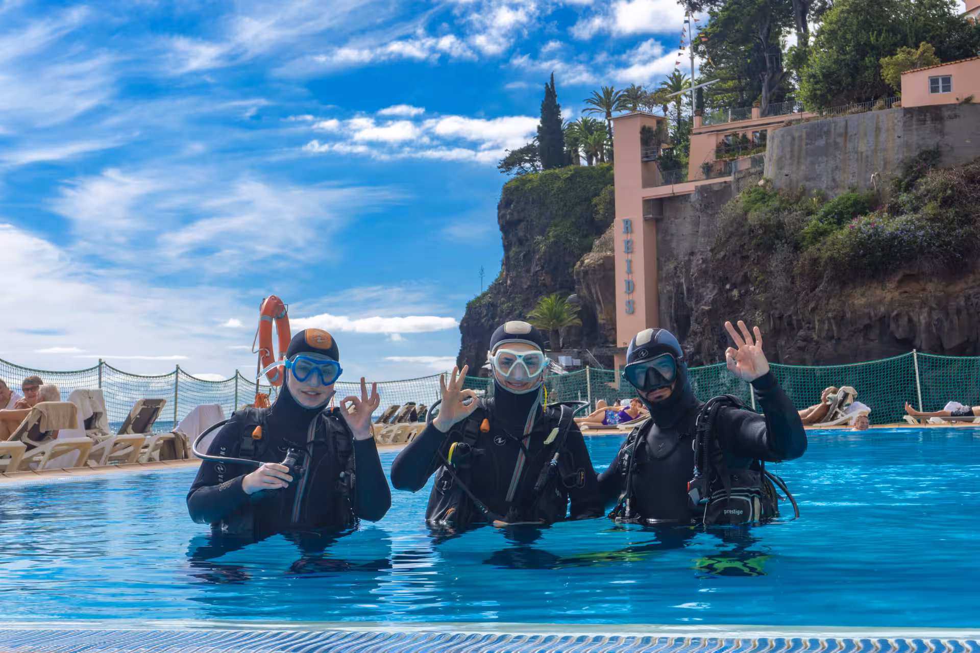Three beginner scuba divers in wetsuits giving OK signs in a pool, ready for an exciting underwater experience.