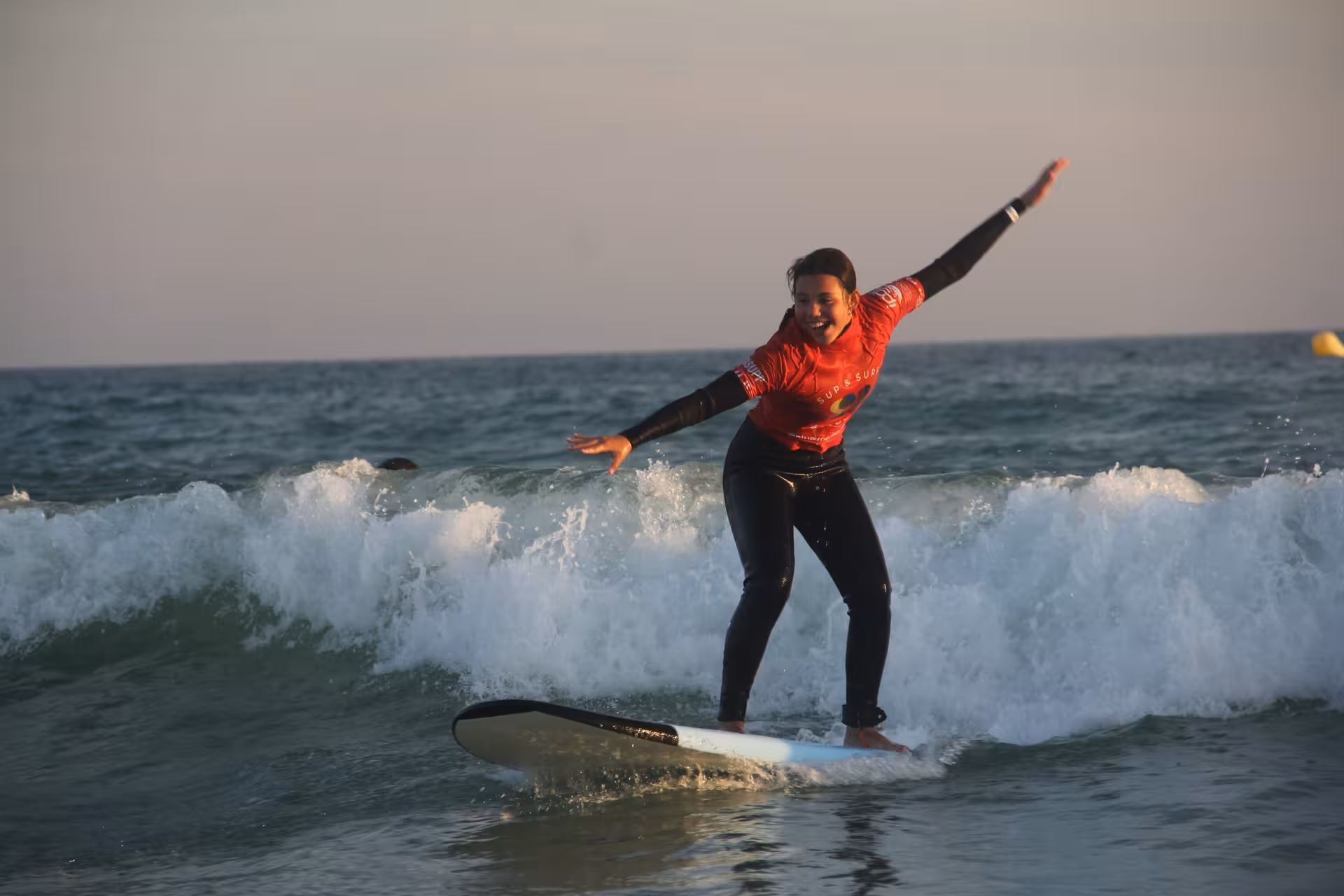 Smiling student riding a beginner-friendly wave in a sunset surf lesson clinic, arms outstretched for balance