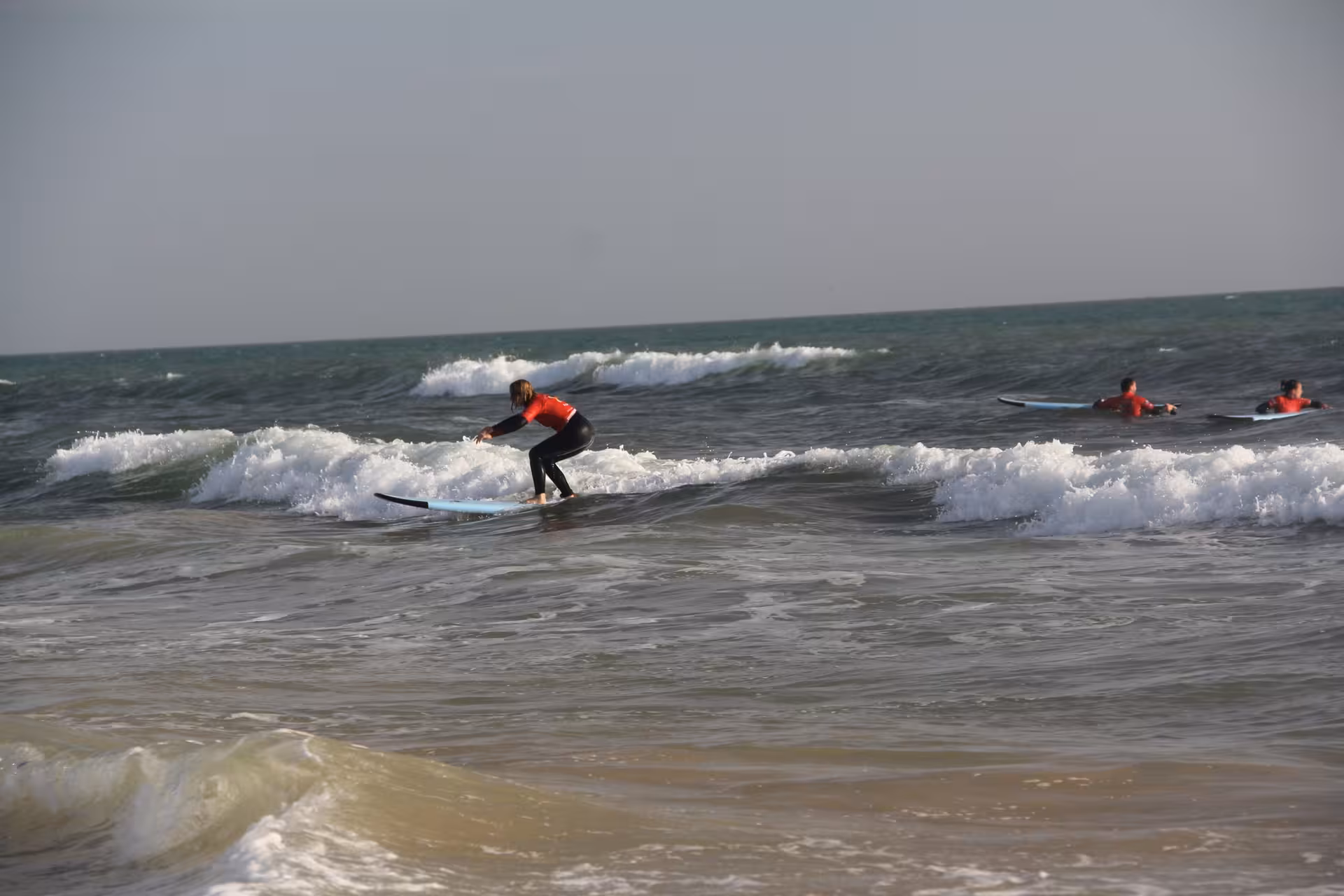 Beginner surfer standing up on a small wave during a 3-hour group surf course with classmates in the background