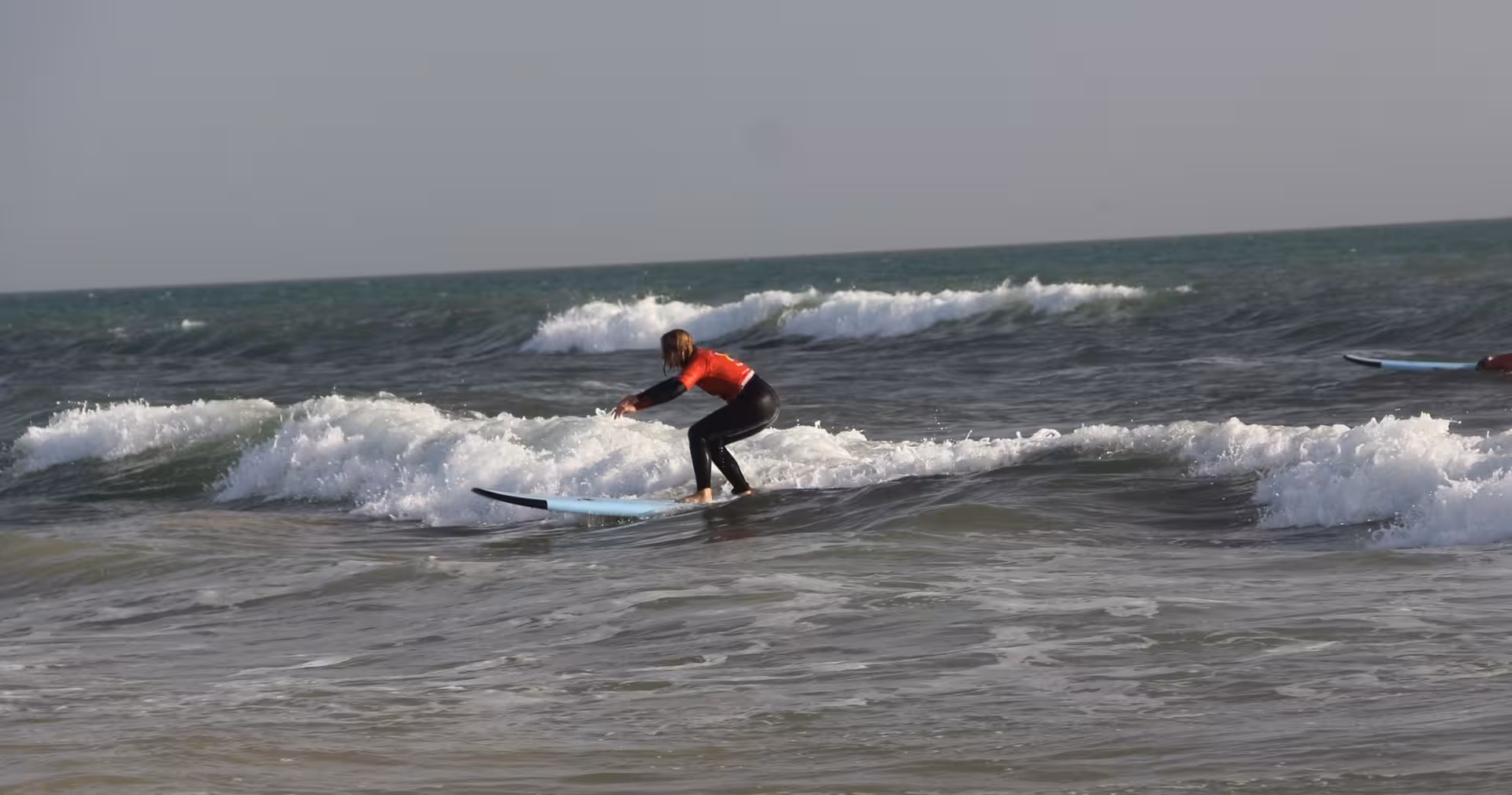 Beginner surfer in red rash guard riding a small wave during a surf lesson clinic on a sunny Atlantic beach