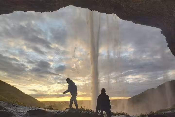 Two silhouetted travelers standing behind Seljalandsfoss waterfall at sunset on Iceland's South Coast