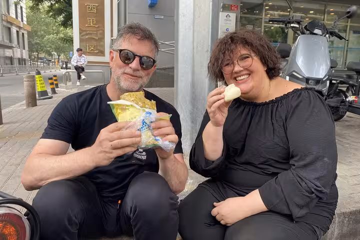 Tourists enjoying traditional Beijing street food during a sidecar tour, savoring local flavors with joy.