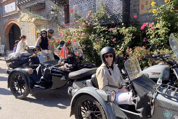 Tourists enjoy a sunny day sidecar tour through historic Beijing, surrounded by vibrant flowers and ancient architecture.