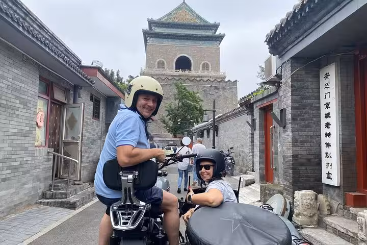 Tourists on a sidecar motorcycle ride through Beijing hutongs with the historic Bell Tower in the background.