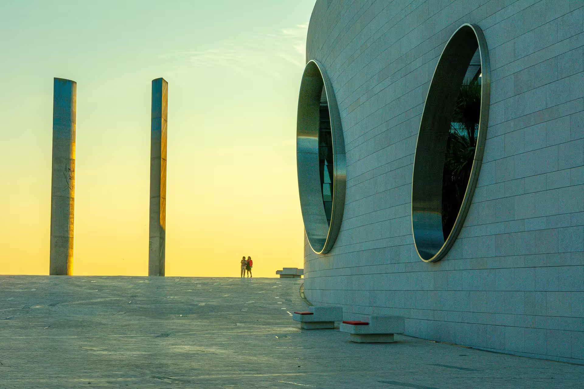 Modern architecture and two pillars silhouetted against a vibrant sunset in Belém, Lisbon, ideal for photography walks.