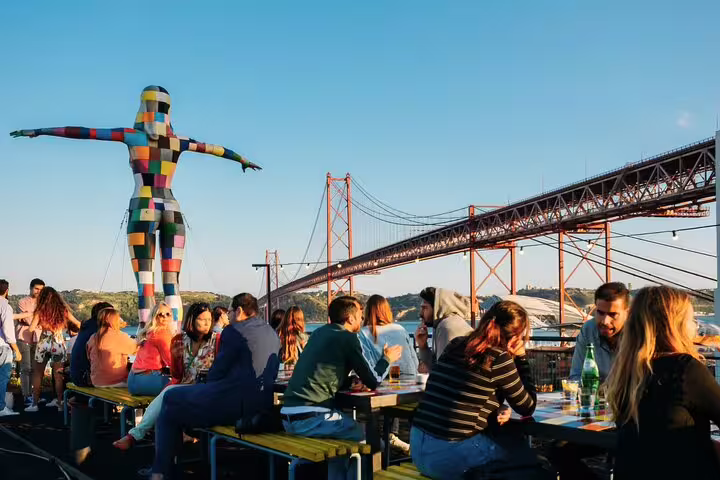 Visitors enjoying a vibrant outdoor dining experience by the iconic 25 de Abril Bridge in Belem, Lisbon, during a private day trip.