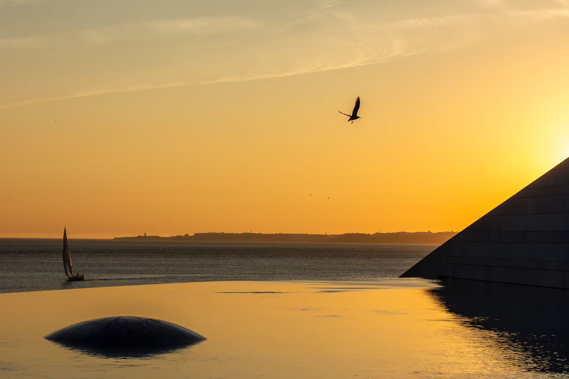 Golden sunset over the Tagus River in Belém, Lisbon, with a sailboat and bird silhouetted against the vibrant sky.