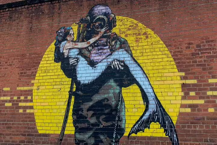 Street art mural of a deep-sea diver carrying a mermaid against a yellow circle on brick wall