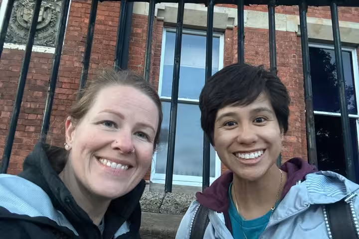 Two smiling women taking a selfie in front of a red brick building with iron gates in Belfast