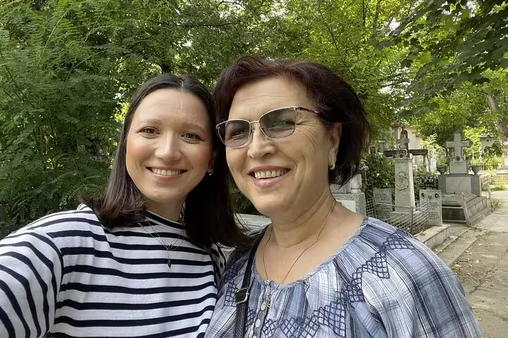 Two women smiling at Bellu Cemetery Bucharest surrounded by green trees and historic tombstones