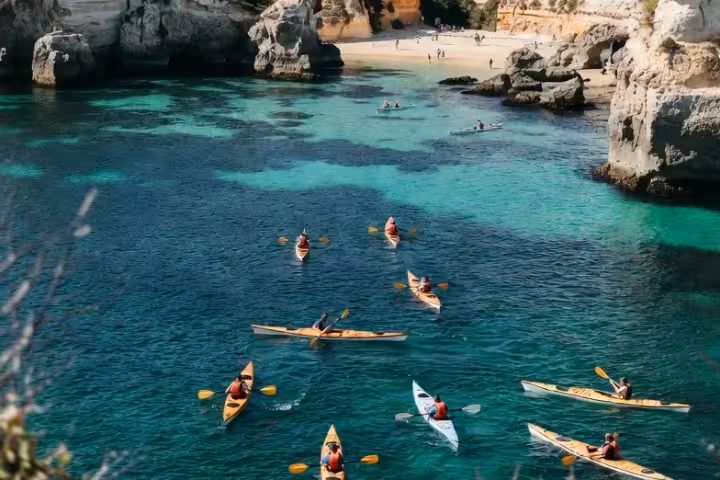 Group of kayakers paddling through crystal-clear turquoise waters near Benagil Beach with stunning limestone cliffs and sandy shore in the background.