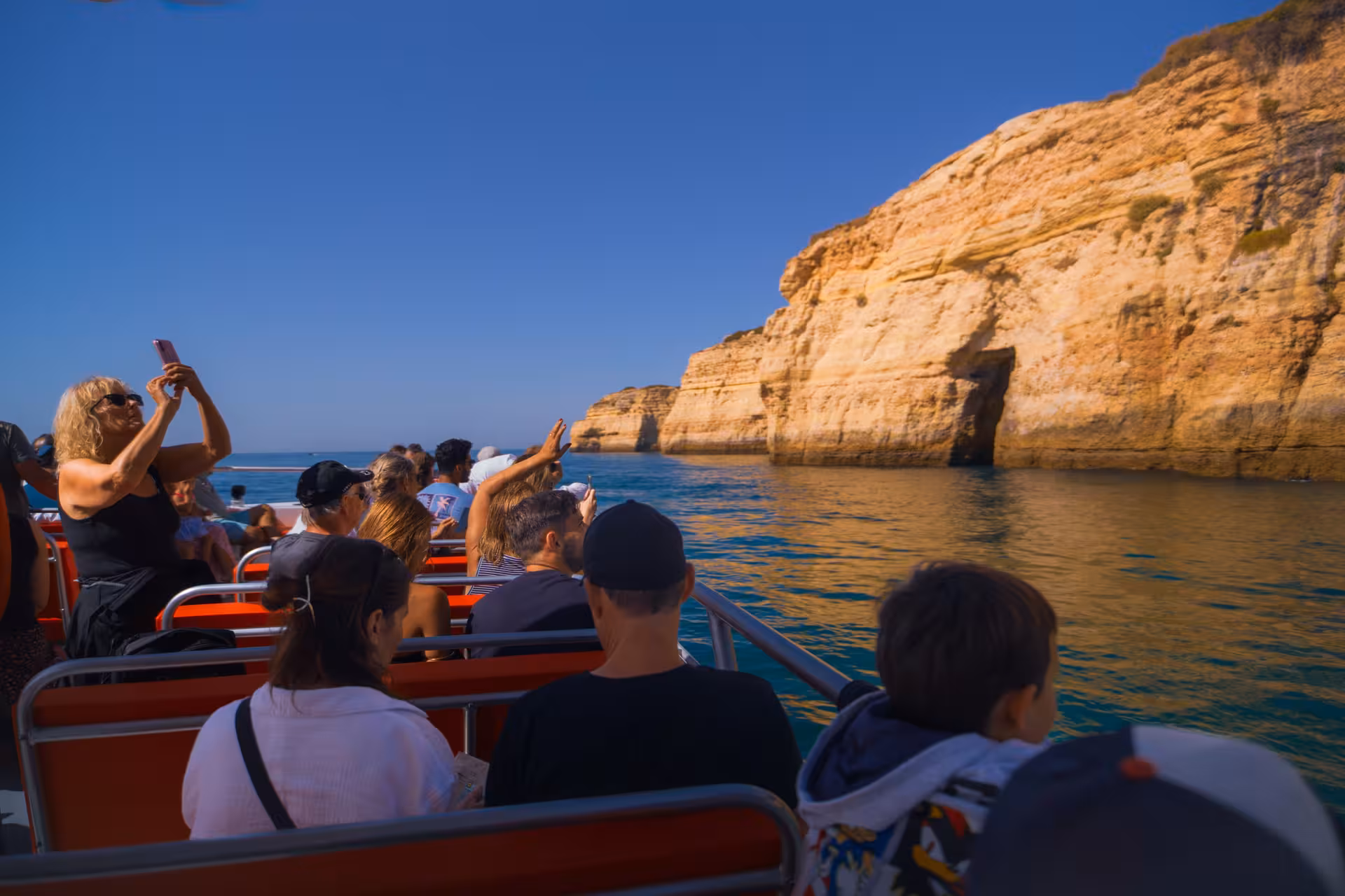 Tourists on a red boat photographing stunning Algarve sea caves and golden limestone cliffs on a guided boat tour