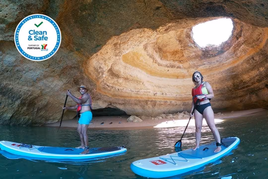 Two friends stand paddling SUP boards by sandy beach inside Benagil Cave on guided Algarve Portugal paddleboard experience
