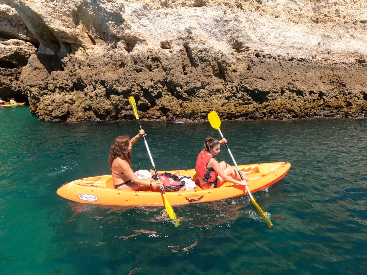 Two women paddle an orange kayak along the scenic rocky coastline of Benagil, enjoying a sunny Algarve day.