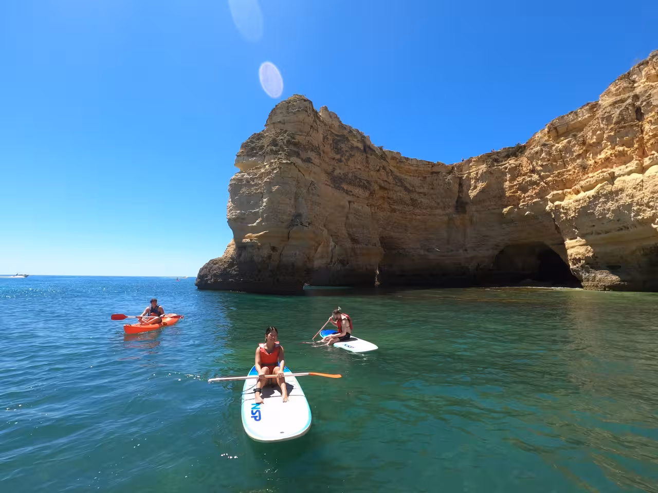 Group of kayakers and paddleboarders enjoy the clear waters and dramatic cliffs of Benagil under the blue sky.