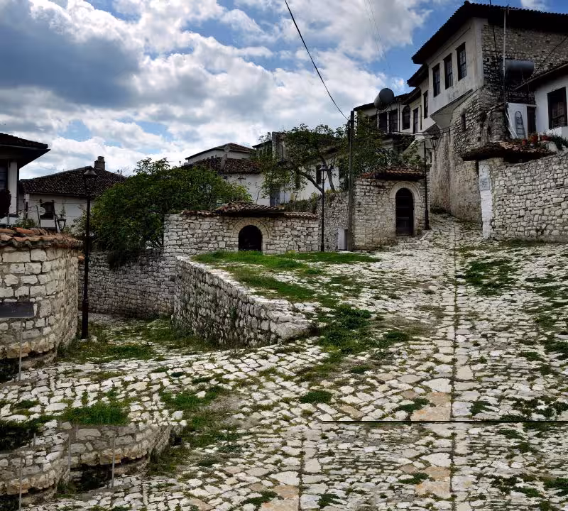 Cobblestone path and traditional stone architecture in Berat, Albania, with lush greenery and cloudy skies.