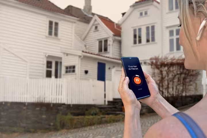 Woman holding smartphone with audio tour app in front of white wooden houses in Bergen, Norway