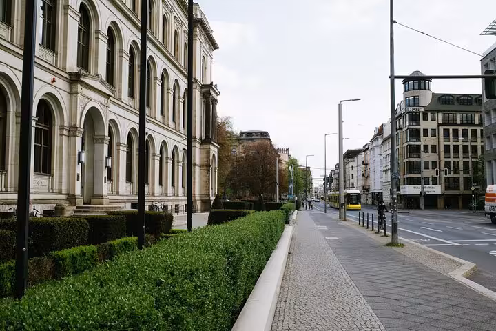 Historic Berlin street with grand neoclassical building, hedges, and yellow tram during Cold War walking tour
