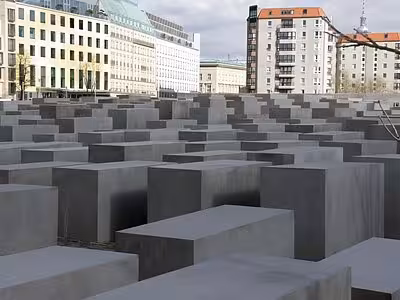 Rows of grey concrete stelae at the Holocaust Memorial in Berlin with city buildings in background