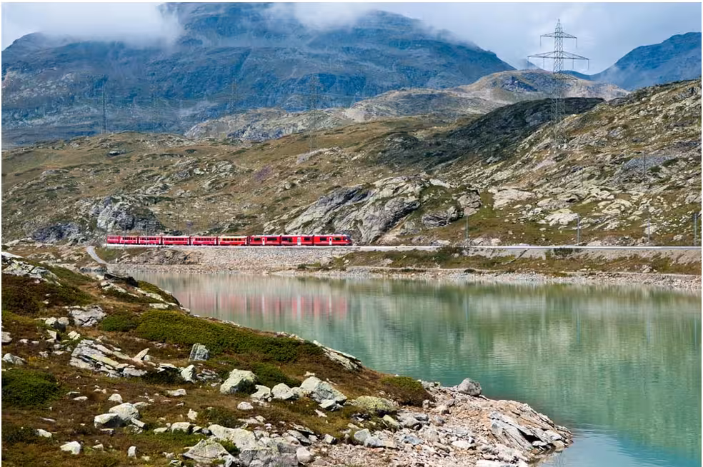 Bernina Red Train passes by a serene alpine lake and rugged mountain terrain, showcasing stunning natural beauty.