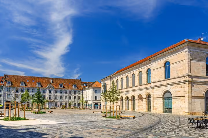 Sunny cobblestone square in Besançon with historic limestone buildings and red rooftops under blue sky