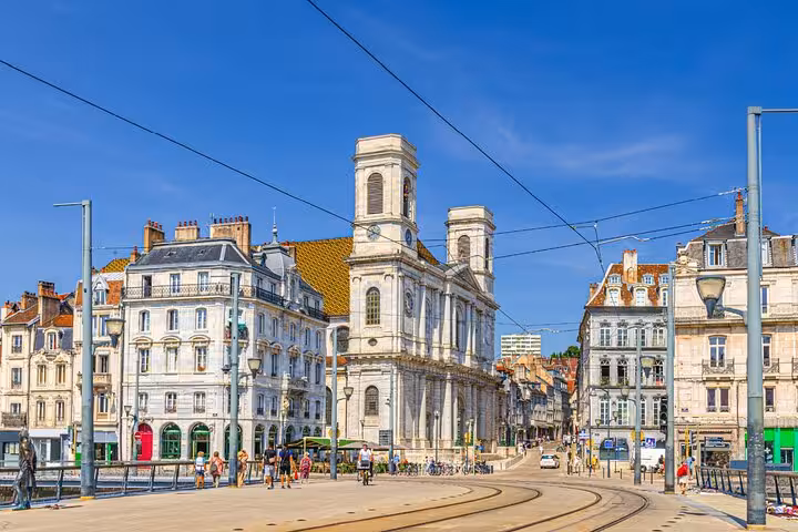 Besancon city square with historic white buildings, church towers, tram tracks, and pedestrians on a sunny day