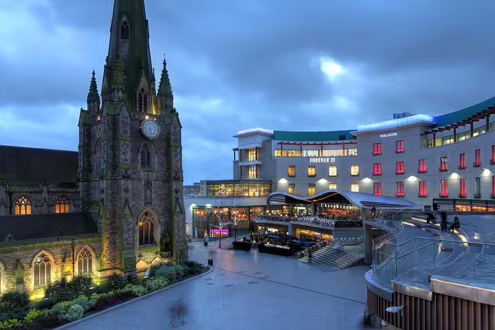 Gothic church spire beside illuminated Forever 21 shopping centre in Birmingham at dusk