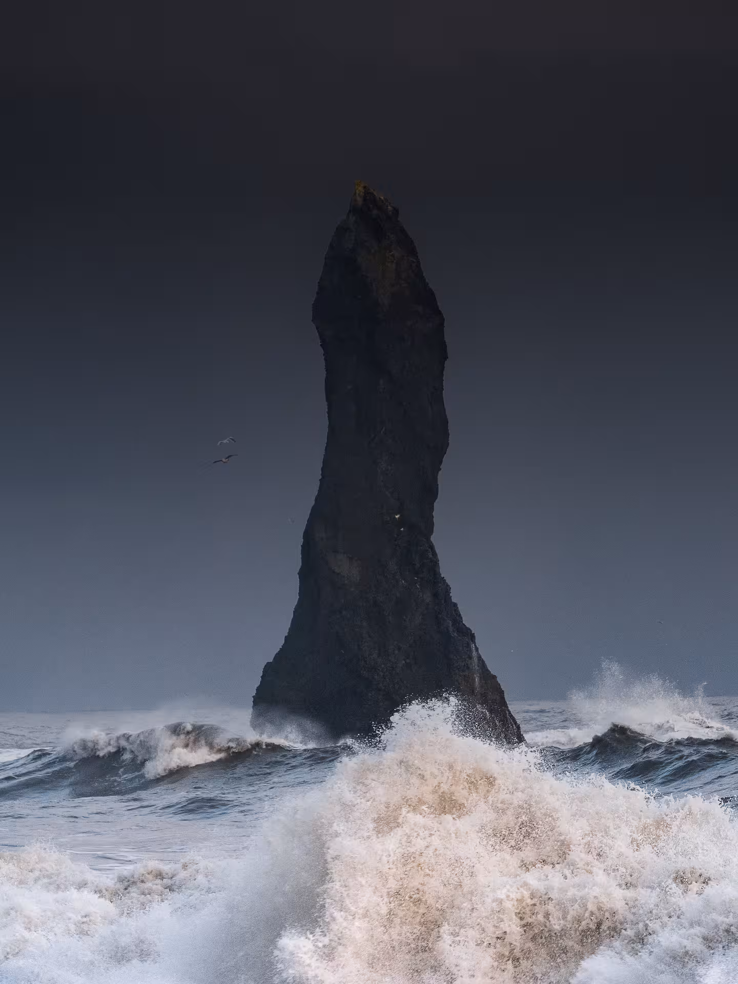 Tall black basalt sea stack rising from crashing waves on Iceland's south coast under stormy skies