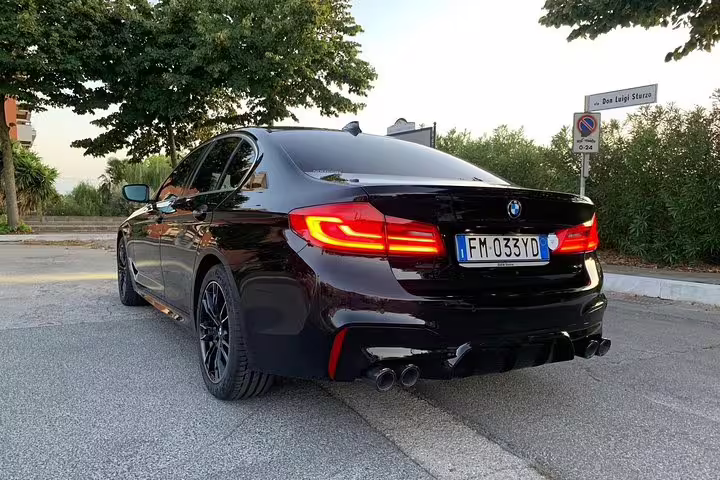 Black BMW sedan rear view with red tail lights parked near trees, Italian license plate FM033YD