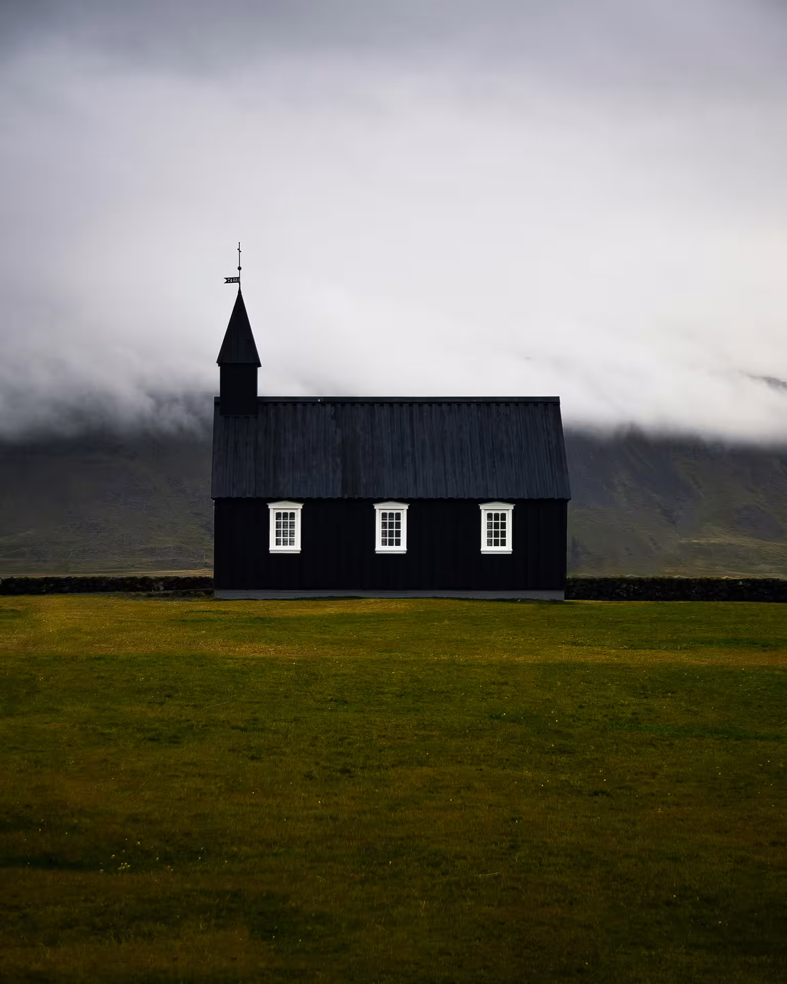 Black wooden church with white windows on green meadow, misty mountains behind, Snaefellsnes Peninsula Iceland