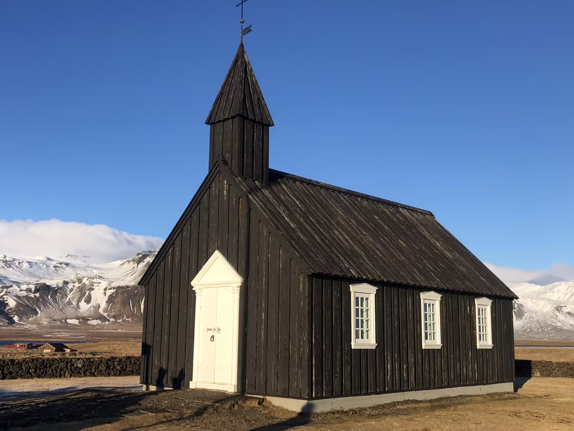 Black wooden church with white door and windows against snow-capped mountains on Snaefellsnes Peninsula, Iceland
