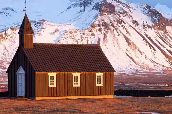 Black wooden church with glowing windows against snow-capped mountain in Snaefellsnes Peninsula, Iceland