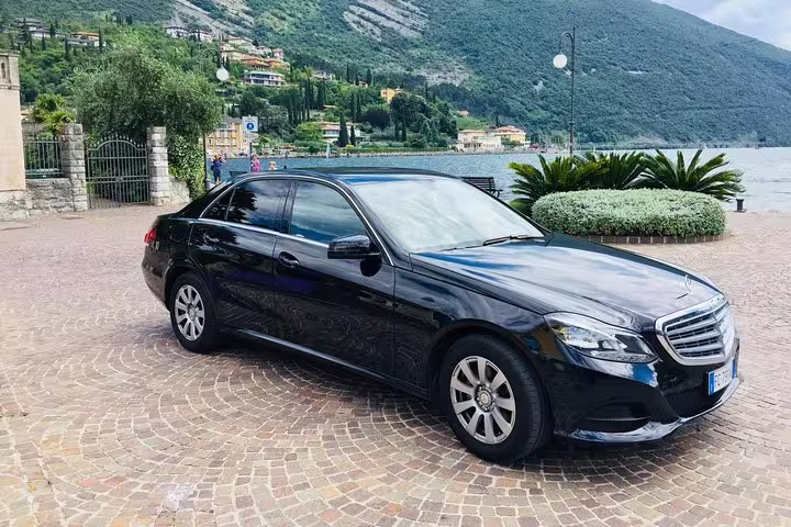 Black Mercedes-Benz sedan parked on cobblestone plaza by lakeside with mountain backdrop