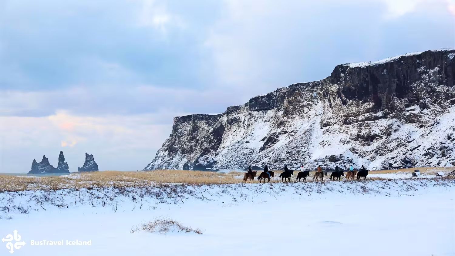 Horseback riders on snowy black sand beach with Reynisdrangar sea stacks and snow-covered cliffs, Iceland winter