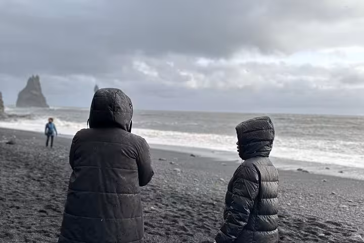 Two visitors in black hooded jackets on Iceland's black sand beach with dramatic sea stacks in background