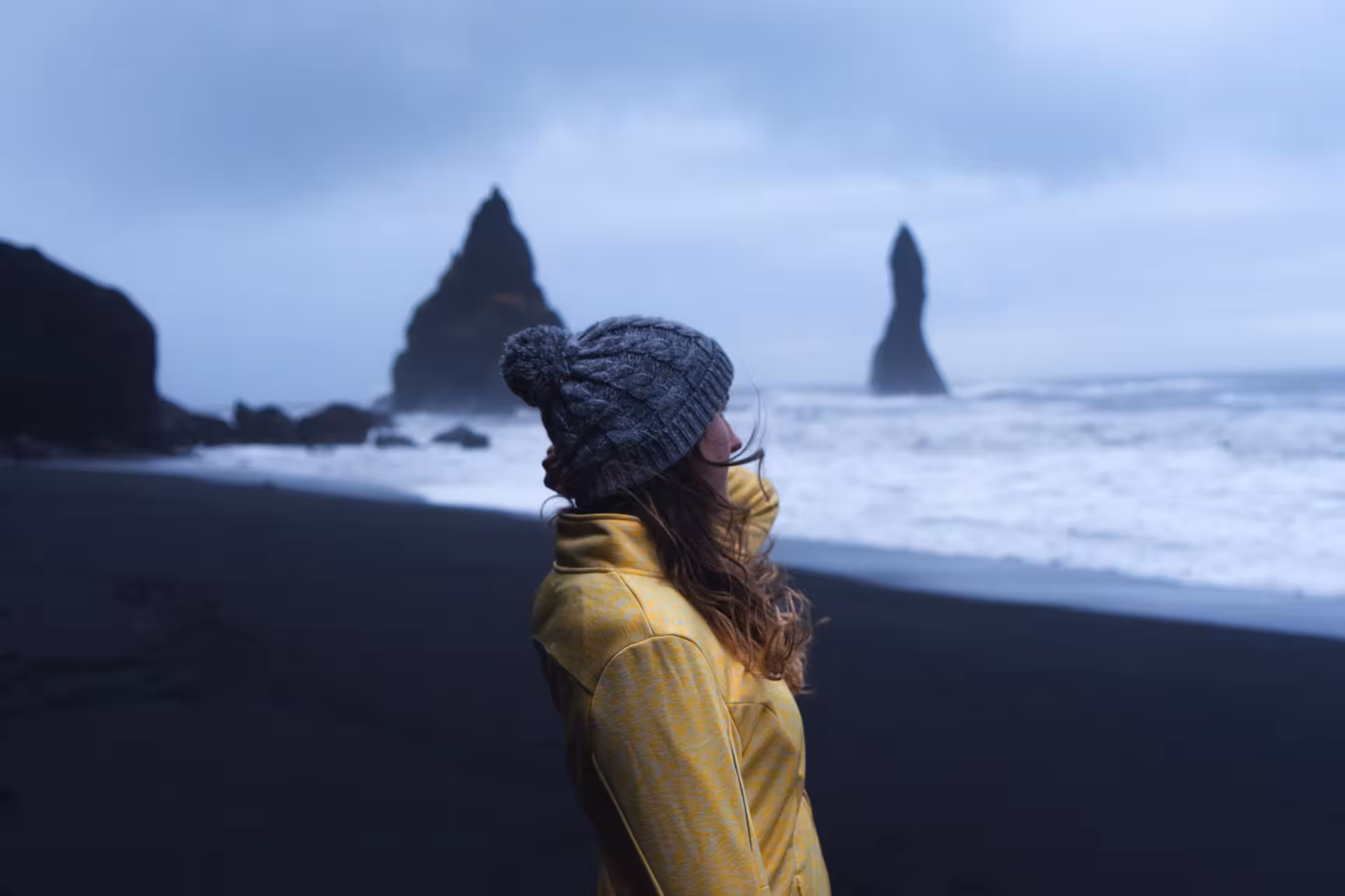 Woman in yellow jacket and grey beanie on Iceland's black sand beach with dramatic sea stacks in background