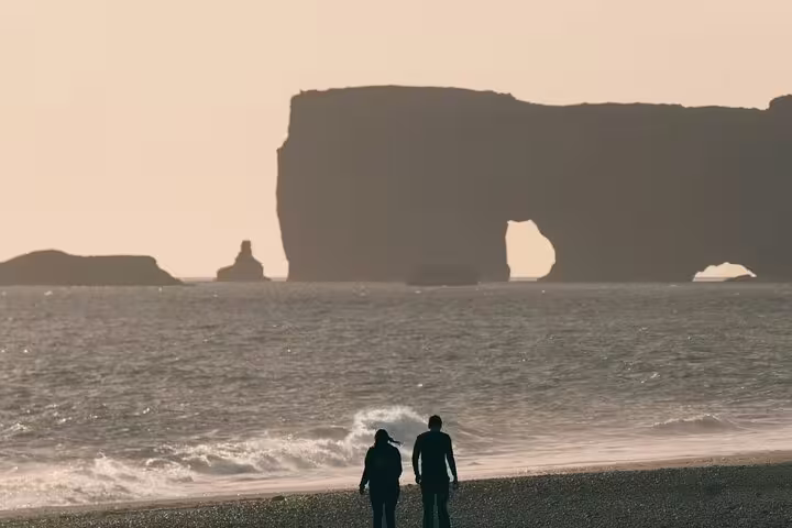 Two visitors standing on Iceland's black sand beach with dramatic coastal rock arch formations at sunset