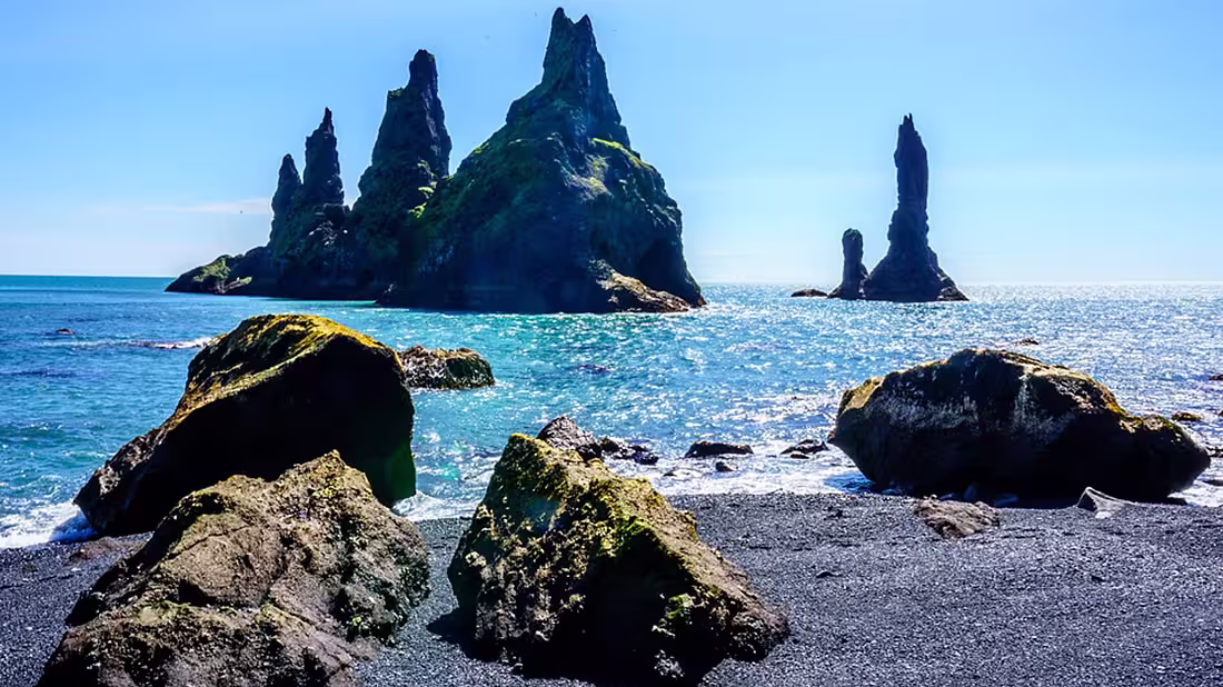 A black sand beach with large rocks in the foreground and island in the background