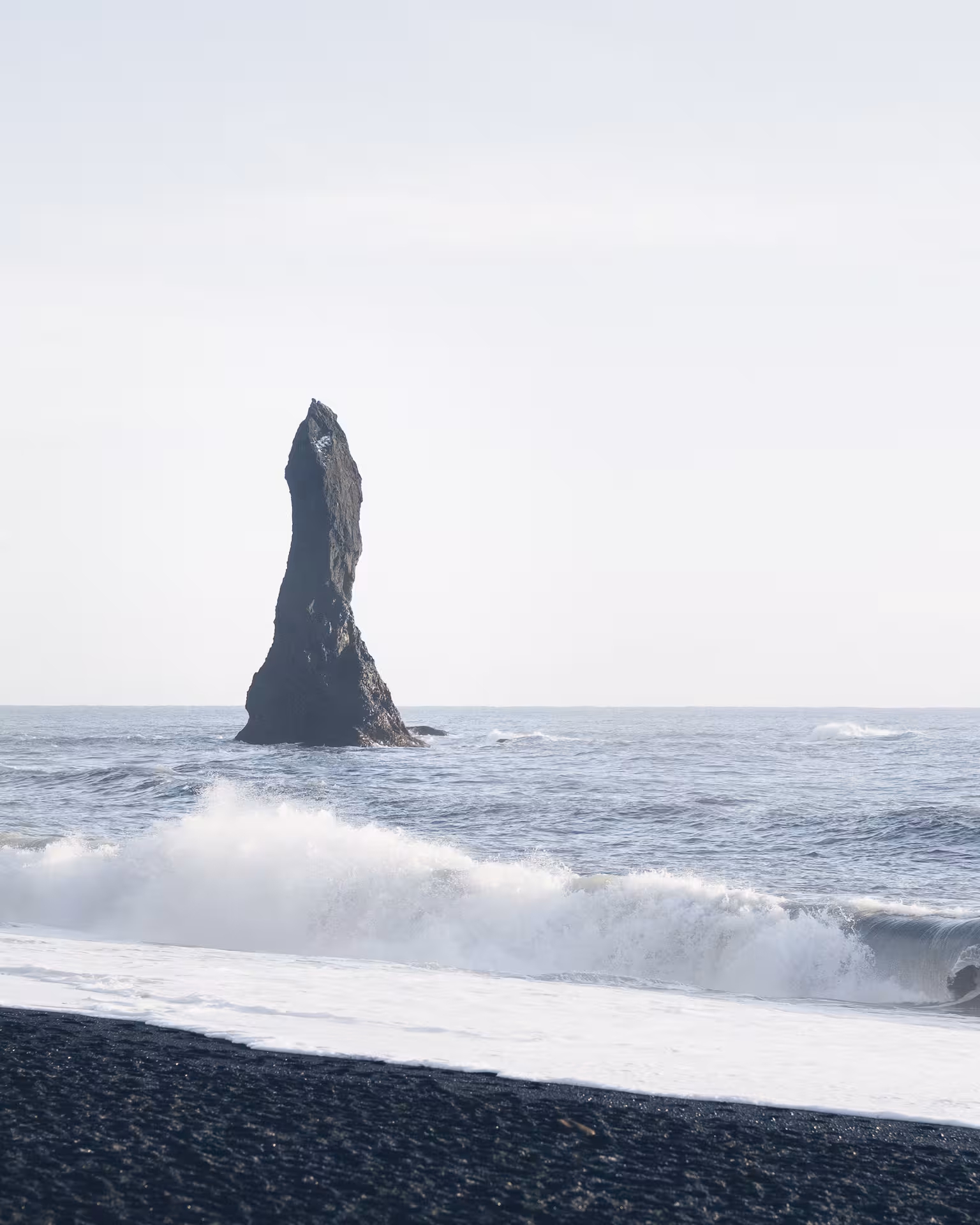 Tall volcanic rock sea stack rising from crashing waves on Iceland's black sand beach