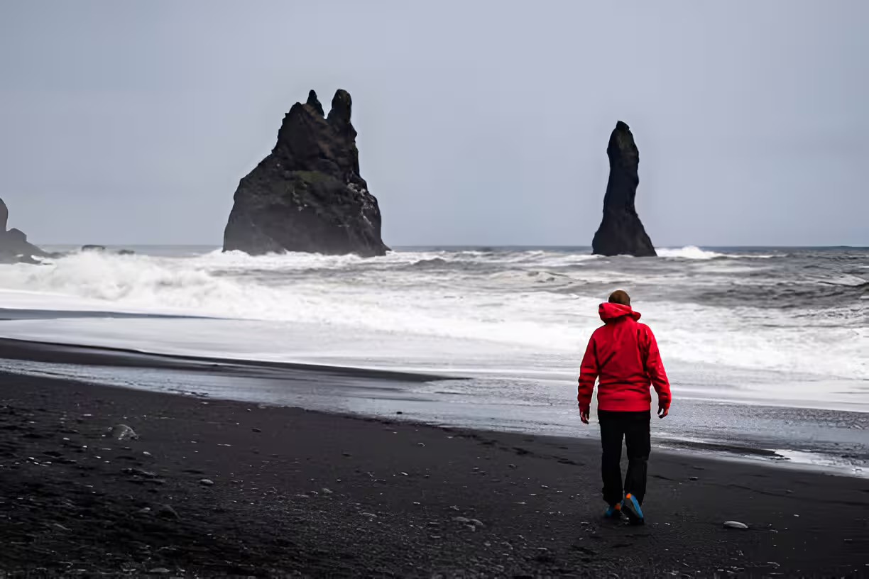 Person in red jacket walking on black sand beach with dramatic sea stacks in Iceland