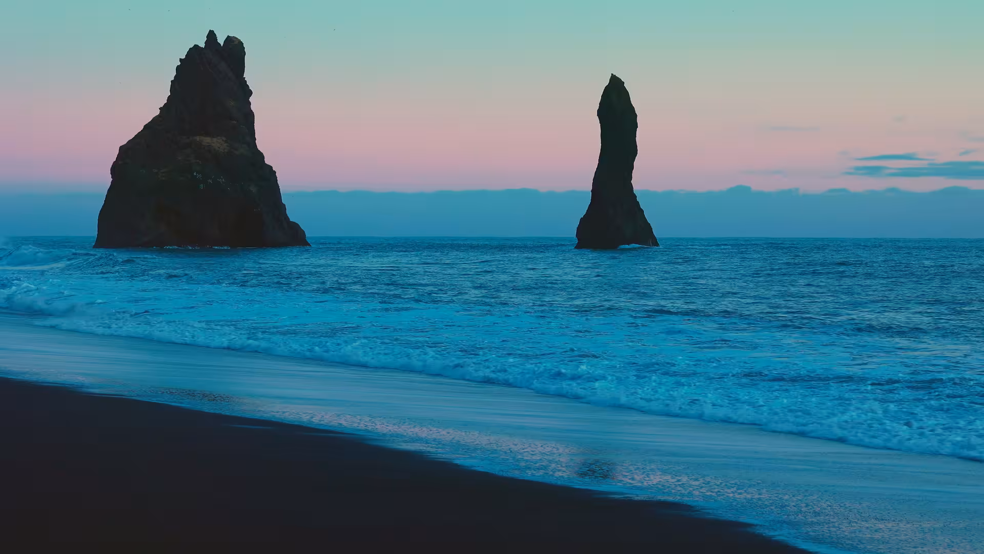 Black sand beach with dramatic sea stacks rising from turquoise ocean under a pink and teal twilight sky