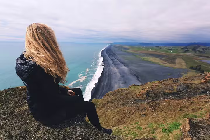 Woman sitting on cliff overlooking Iceland's black sand beach and turquoise ocean on south coast tour