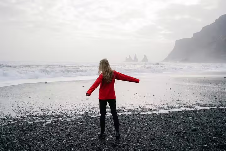 Woman in red jacket standing on Iceland's black sand beach with dramatic waves and sea stacks