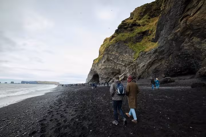 Tourists walking along Iceland's black sand beach beside towering volcanic cliffs on the South Coast