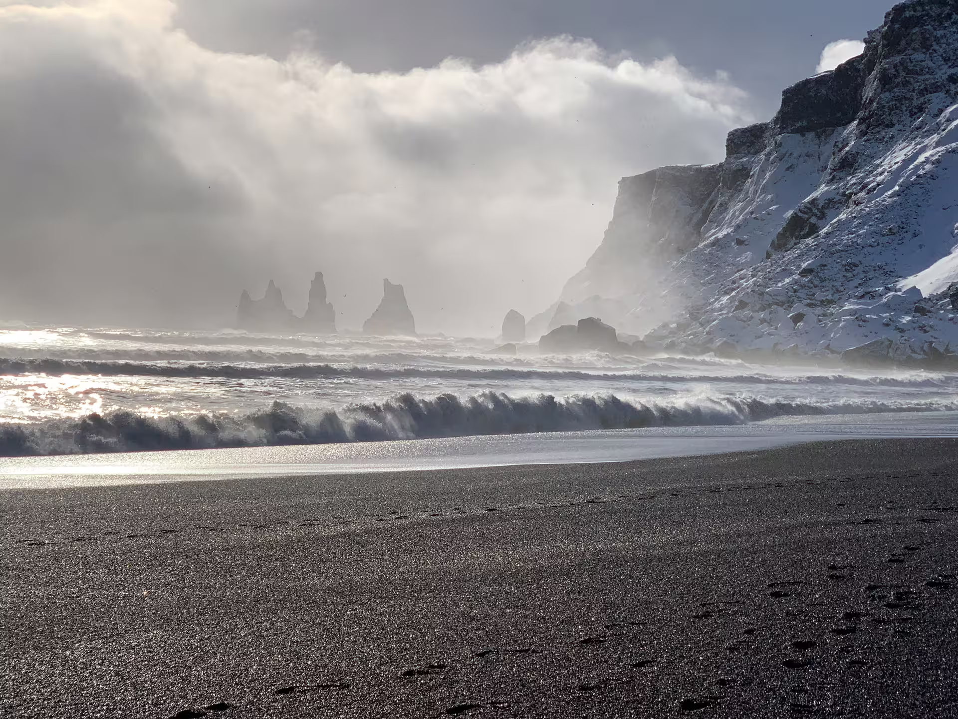 Black sand beach with dramatic sea stacks and snow-covered cliffs during stormy winter conditions in Iceland