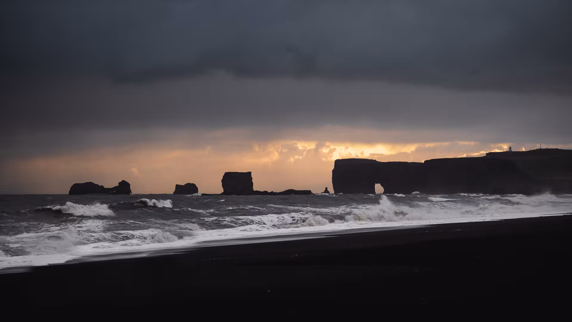 Black sand beach with dramatic waves and volcanic rock formations silhouetted against stormy Iceland sunset sky