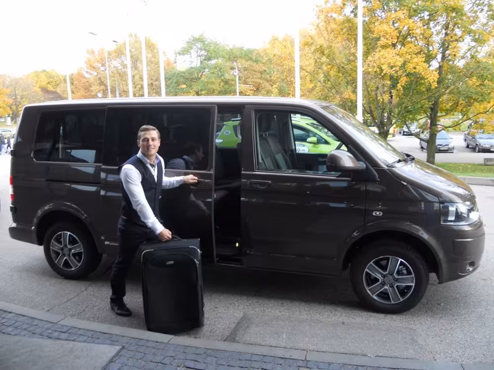 Driver standing beside black Volkswagen Transporter van with luggage for private airport transfer