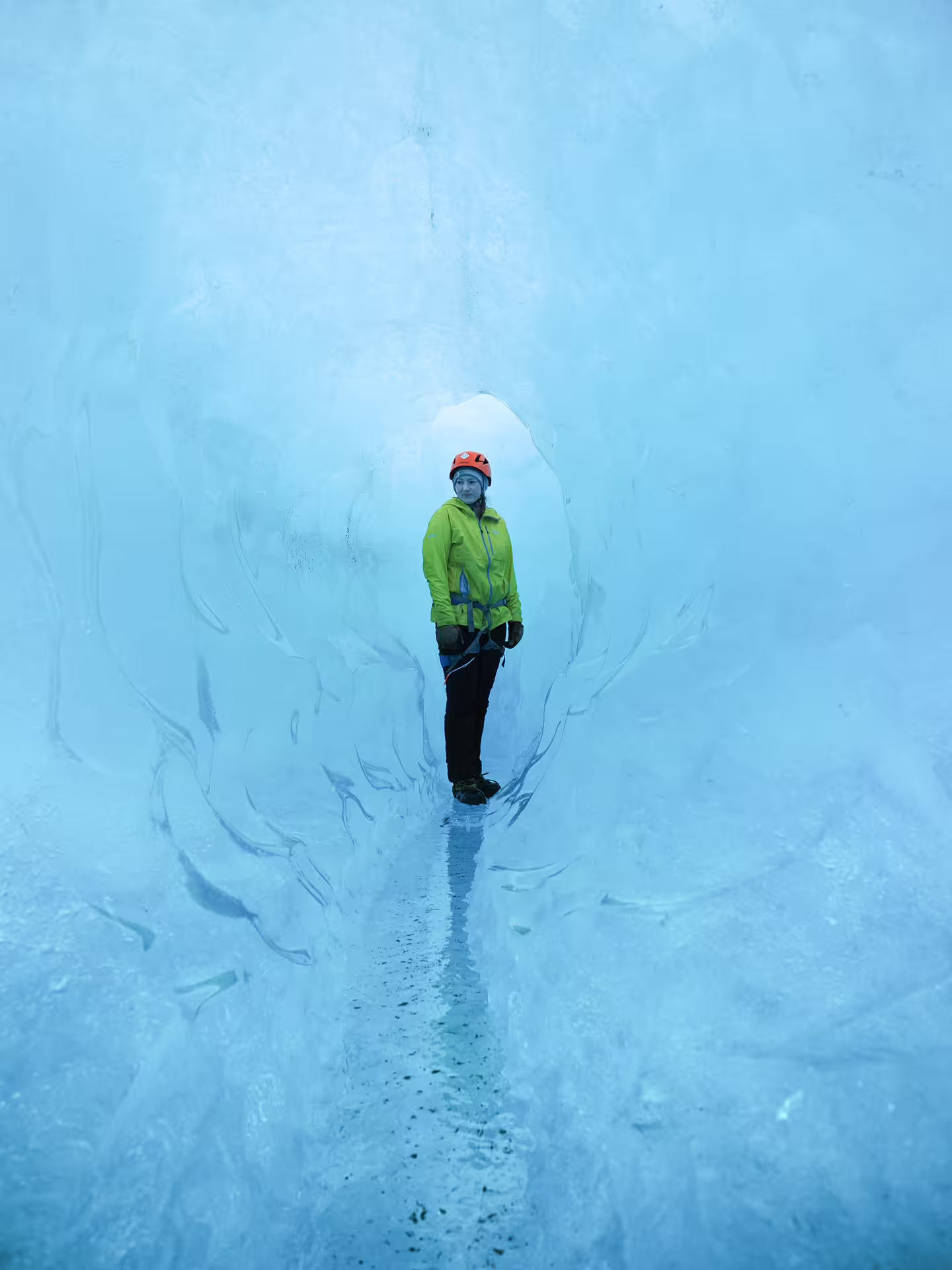 A hiker in a green jacket and orange helmet stands inside a stunning blue glacier ice cave tunnel on a winter adventure tour.