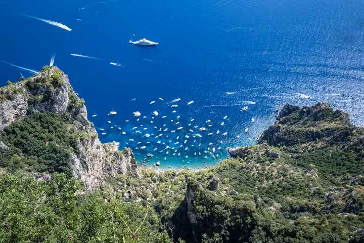 Aerial view of boats near the iconic Blue Grotto and rugged cliffs of Faraglioni, Capri, under a clear blue sky.