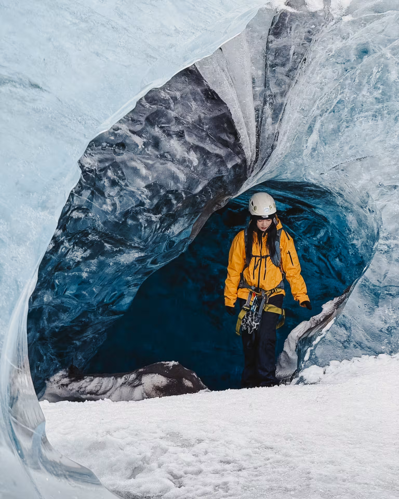 Gear-equipped explorer in yellow jacket exiting a stunning blue ice cave entrance on Iceland glacier tour